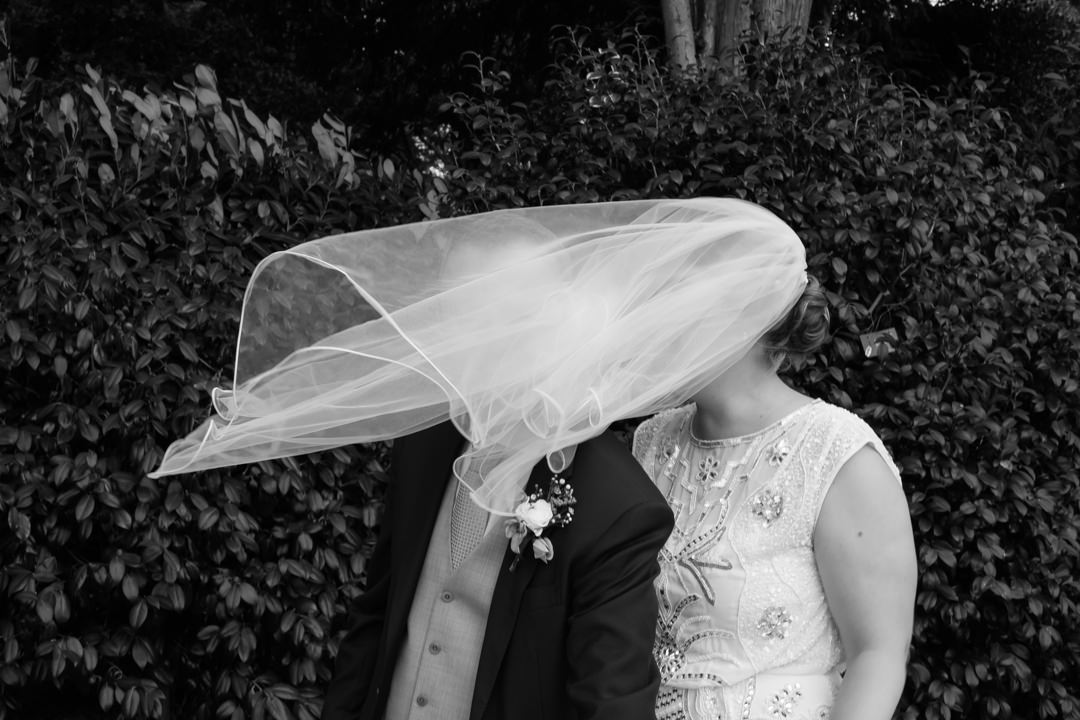 Brides veil blows over the face of the groom as they wait to cut their cake. Black and white photo