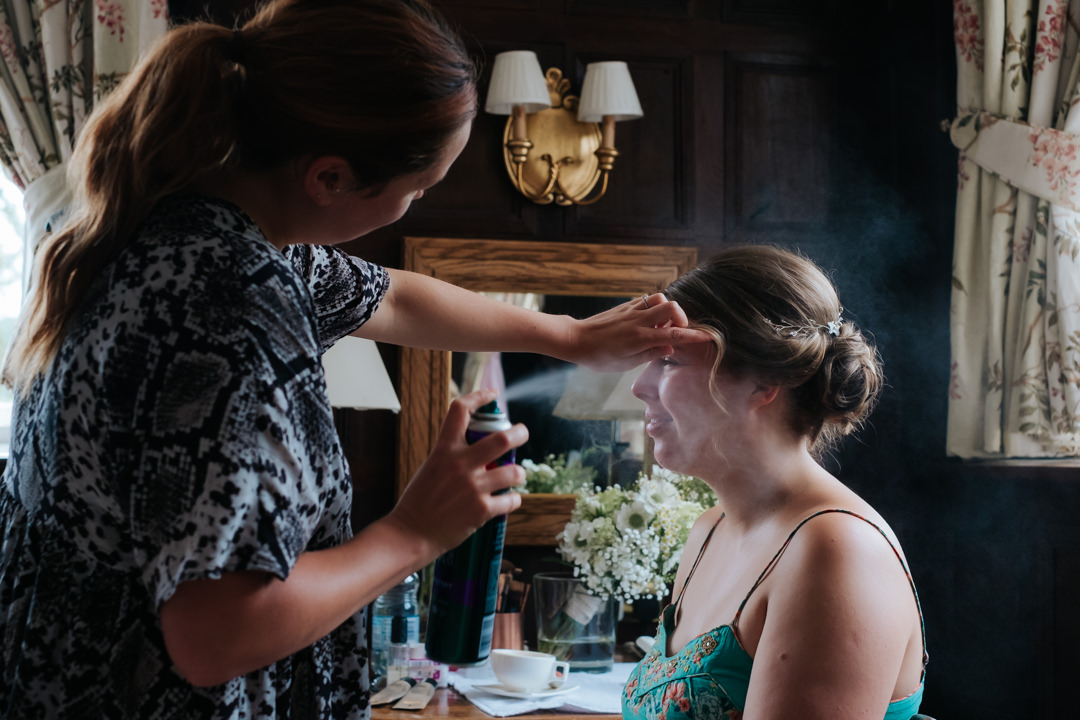 hairdresser sprays hair spray onto beautiful bride in green dress as she gets ready for her wedding in oak panelled room at Gravetye Manor wedding venue