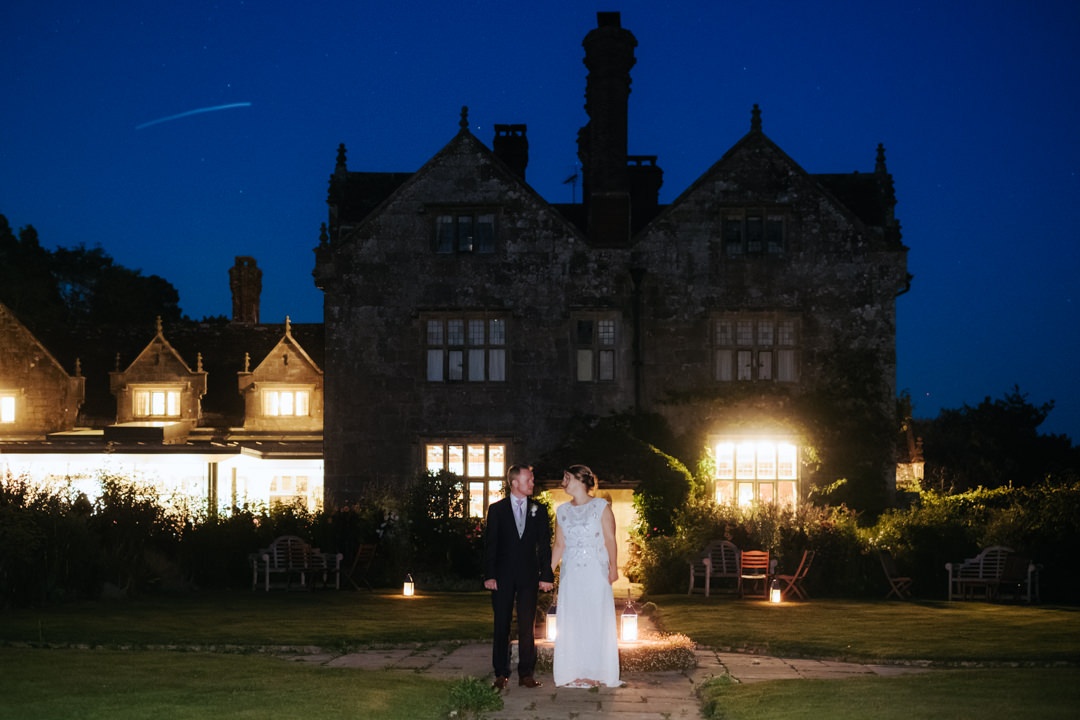 Bride and groom stand in the dark outside the house at Gravetye Manor. backlit photograph showing dep blue night time skies and a shooting star