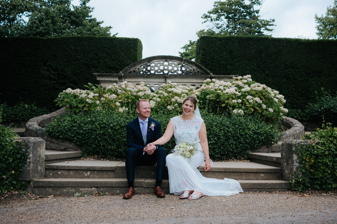 Bride and groom sit together holding hands beneath beautiful cream flowers after their wedding at Nymans Gardens