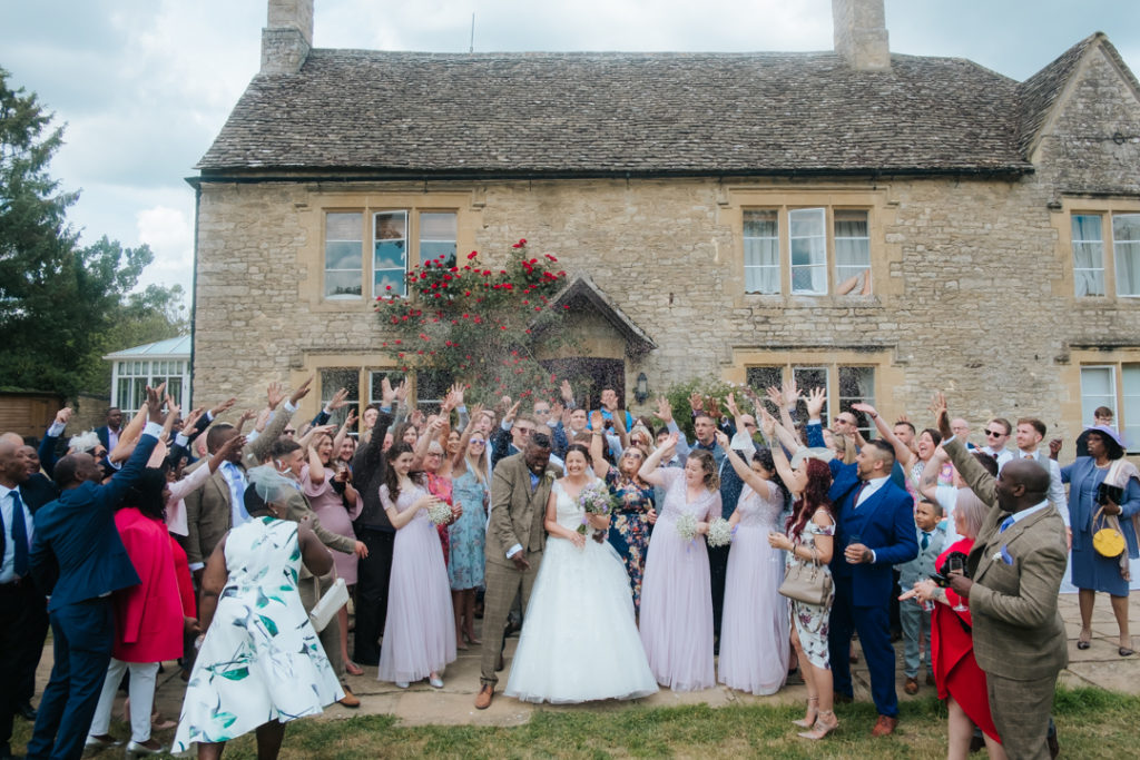 wedding guests throw confetti over the bride and groom, stood in front of the old farmhouse at their Worton Hall wedding