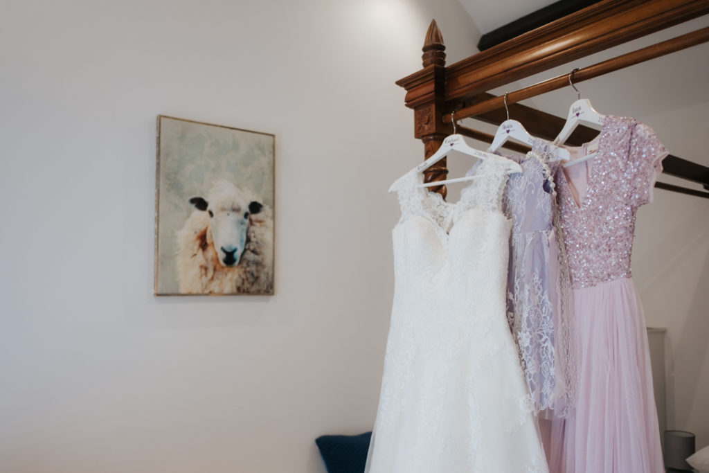 White wedding dress and pink sequinned bridesmaids dress hanging from bed next to sheep painting at Worton Hall wedding venue on the morning of Rachael's African Fusion wedding