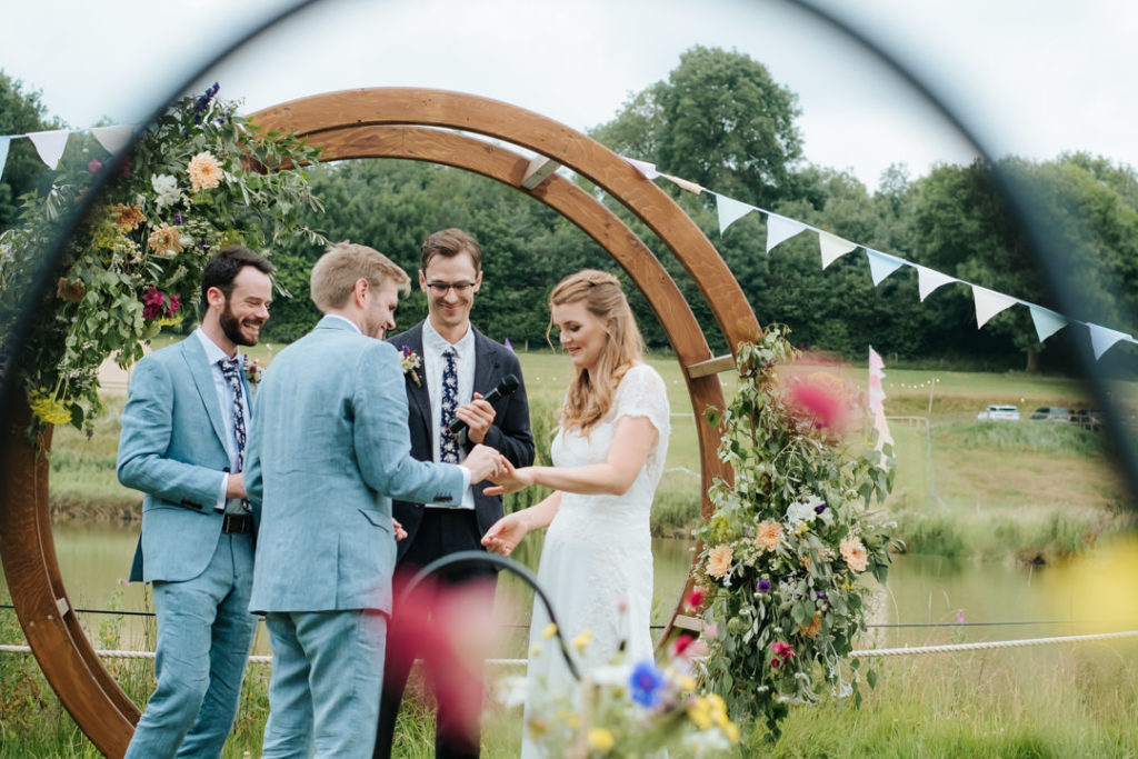 Grooms puts his wedding ring onto the Bride's finger in front of the flower arch at their festival wedding next to the lake at Hadsham Farm Wedding venue in Banbury.