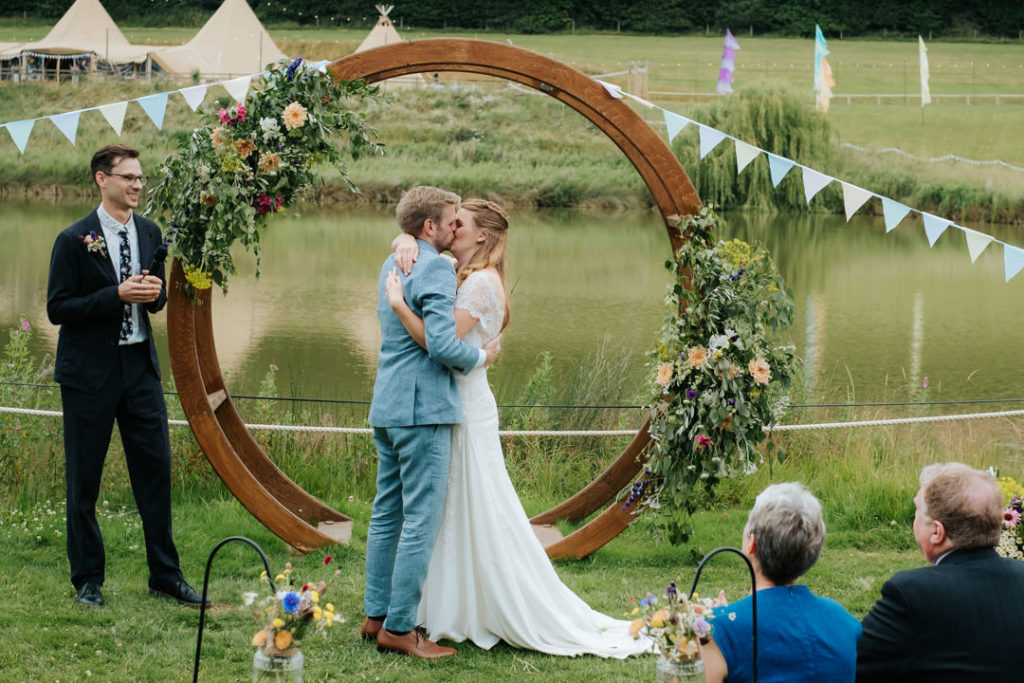 Bride and groom embrace and have their first married kiss in front of the flower arch next to the lake at Hadsham Farm Wedding venue in Banbury.