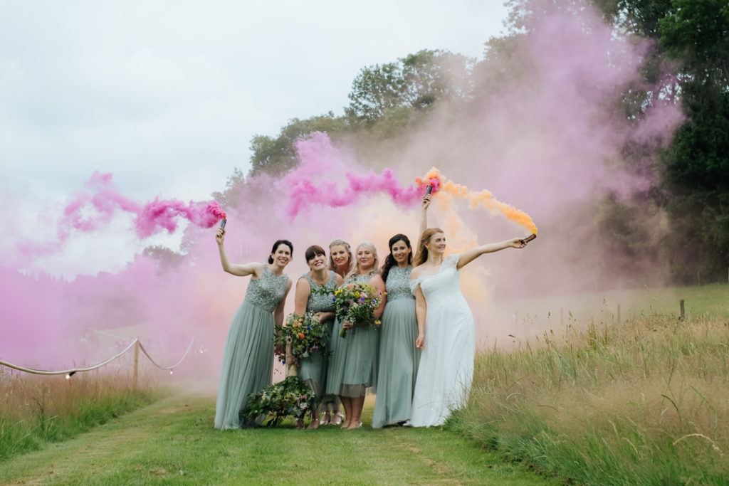 bride and bridesmaids stand together at Hadsham Farm Weddings waving pink and orange smoke bomb flares