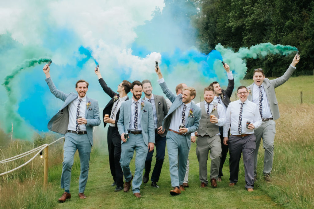 Groom and male ushers walk in the ground of Hadsham Farm Weddings whilst waving blue and green smoke bomb flares