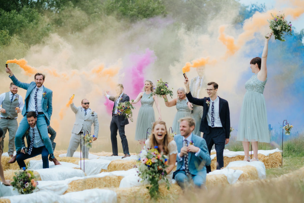 Bride and groom laugh to see their ushers and bridesmaids having fun with coloured smoke bombs in the air after their relaxed wedding ceremony at Hadsham farm weddings. A favourite kind of wedding for charlie Flounders to photograph