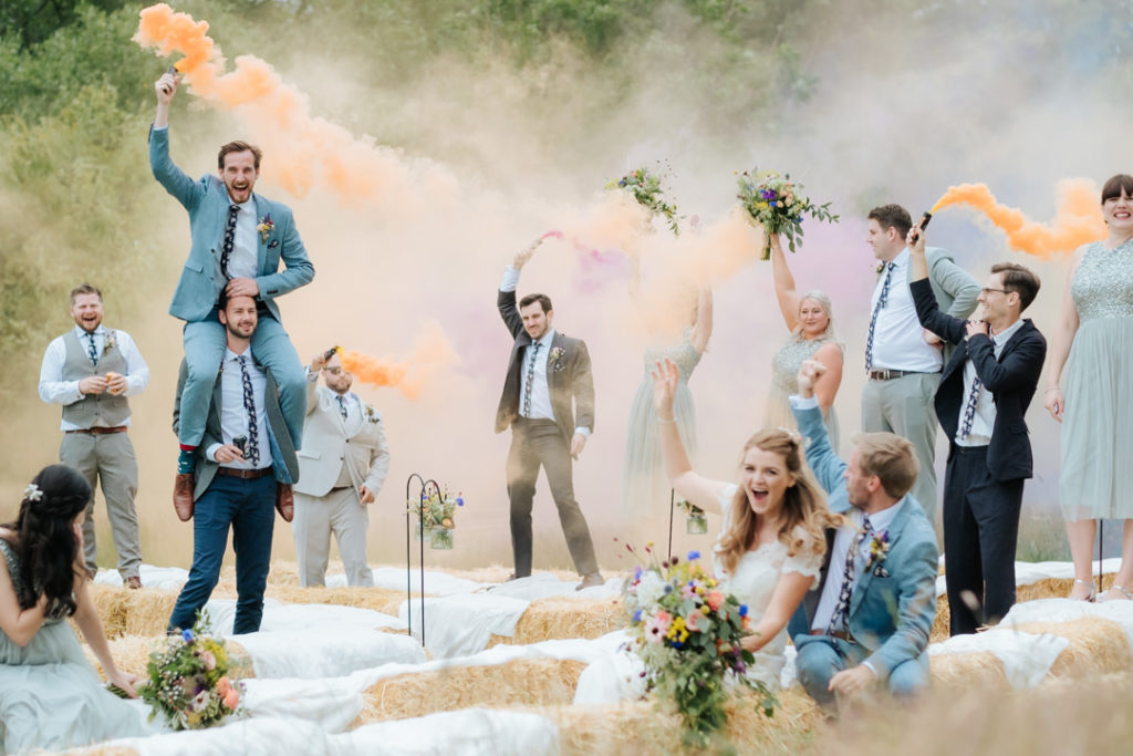 Bride and groom laugh and cheer to see their ushers and bridesmaids being silly and waving coloured smoke bombs in the air after their relaxed wedding ceremony at Hadsham farm weddings. my favourite kind of wedding to photograph