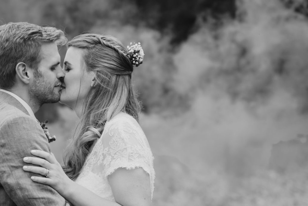 blakc and white photograph of bride and groom kissing after thier wedding at Hadsham Farm weddings in Banbury. Smoke from coloured smoke bombs lingers in the background
