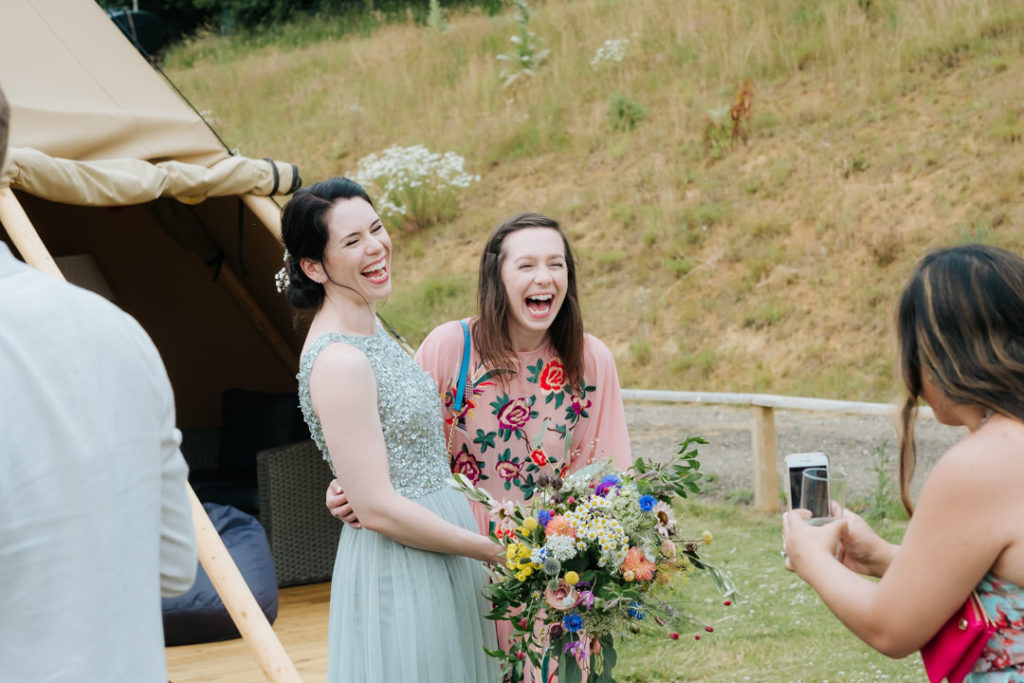 Bridesmaid and her friend laugh holding the bridesmaids bouquet whilst their friend takes a photo of them at Hadsham Farm Weddings