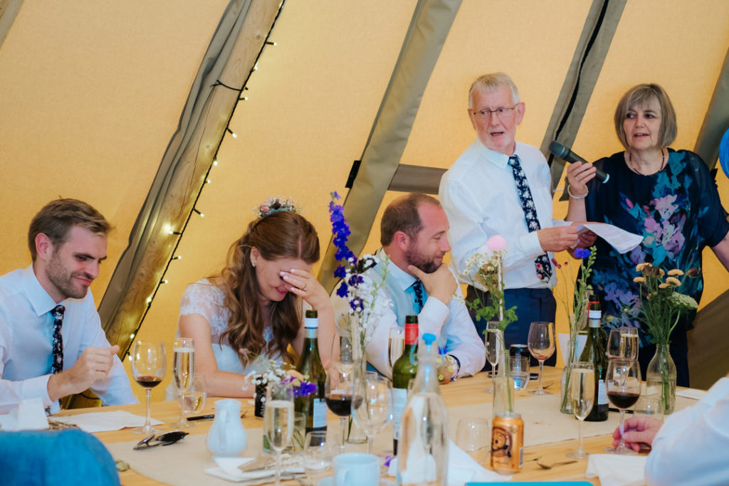 bride holds her head whilst listening to her Dad's wedding speech inside the tipi at Hadsham Farm weddings Branbury. Such a fun festival wedding to photograph