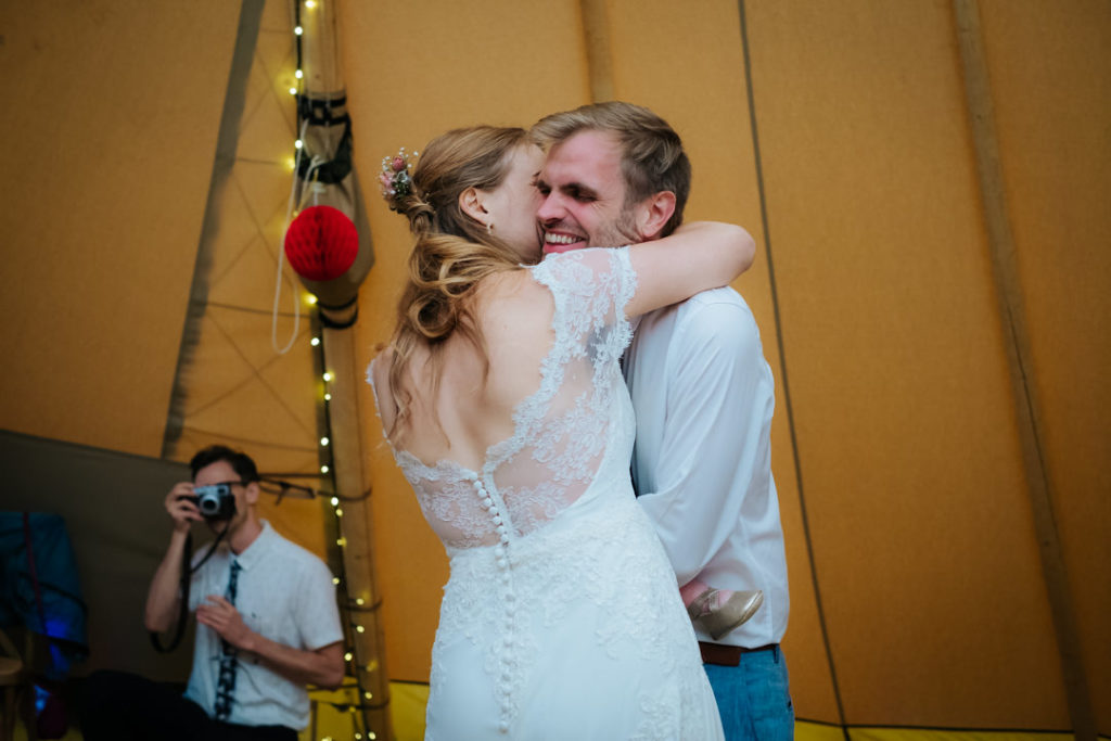 bride and groom embrace as they perform their first dance inside the tipi at Hadsham Farm weddings, Banbury as their friend stands behind them to take a photograph. Such a fun wedding to photograph