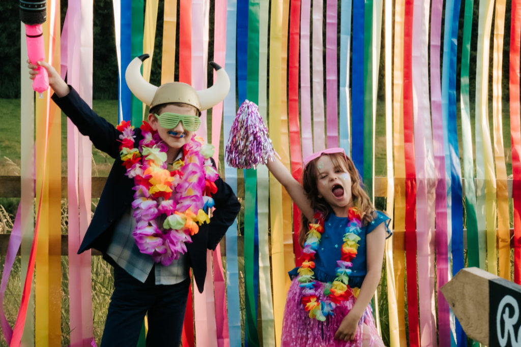 2 children pose with their arms in the air in front of a rainbow photo backdrop with silly hats and glasses on at Hadsham Farm weddings, Banbury