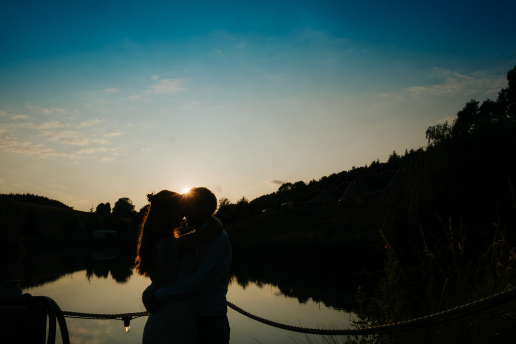 silhouette of bride and groom kissing at sunset in front of the lake at Hadsham Farm weddings, Banbury. the flare of the sun shines through over the couple's head and reflects on the lake.