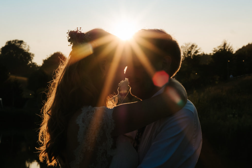 The sun is setting behind a bride and groom who embrace at sunset in front of the lake at Hadsham Farm weddings, Banbury. The flare of the sun shines over the top of the heads of the couple and through her hair.