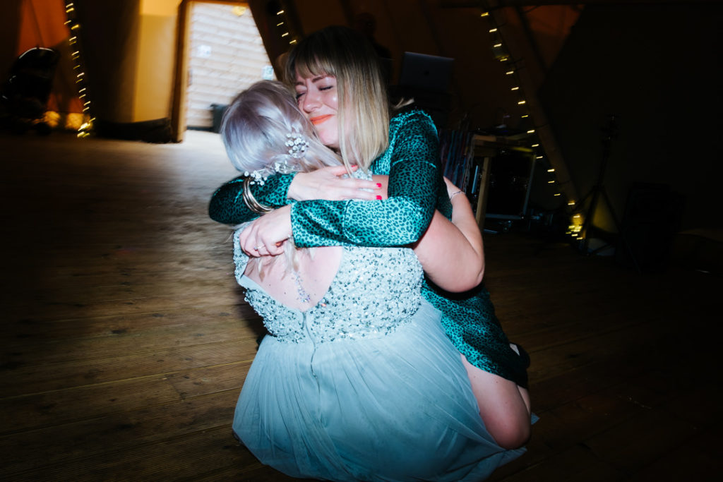 2 female friends hug whilst sitting on the floor having just fallen over whilst dancing inside a tipi at Hadsham farm wedding