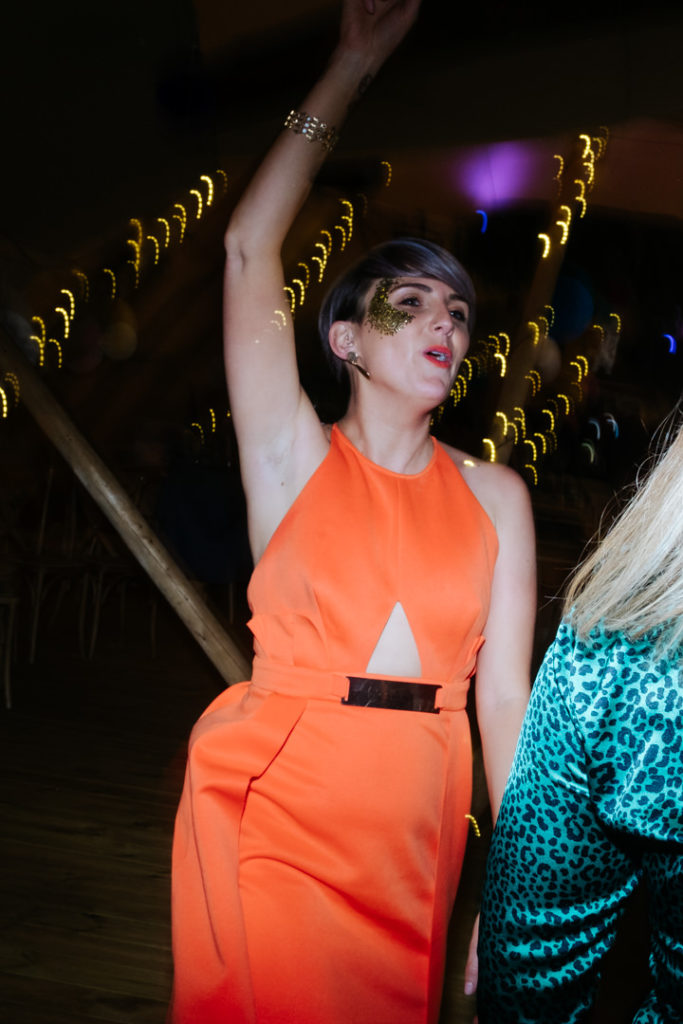 attractive blonde lady wearing orange dress and gold glitter on her face dances with her hands in the air at Hadsham farm tipi wedding. My fun wedding photography style enabled me to catch this.