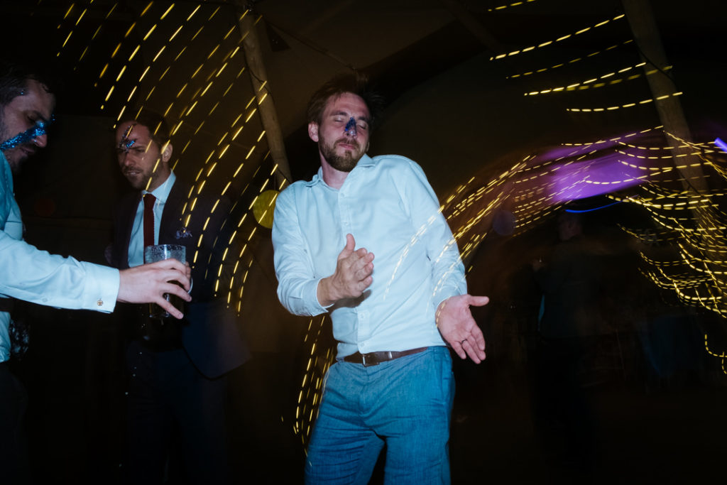 Man wearing white shirt, with glitter on his face dances with his hands out on the dance floor of a tipi at a wedding at Hadsham farm weddings. Light trails can be seen across the image in the backgroun