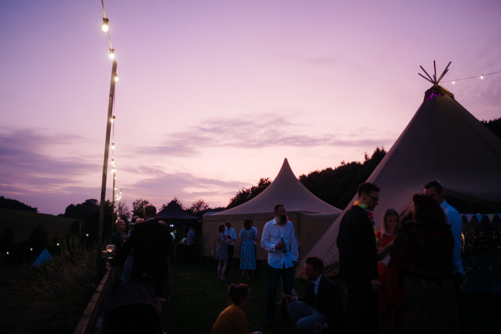 Evening view over the tipi at Hadsham Farm wedding venue as the sun sets. Guests enjoy drinks in the warm summer's evening outside the tipi's under festoon lights and a beautiful purple orange sky