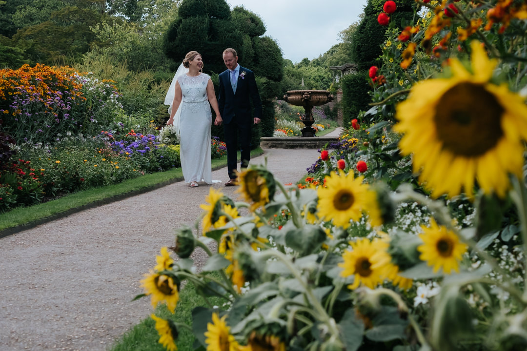 bride and groom walk along the pathway lines with sunflowers and other red and purple flowers whilst holding hands.