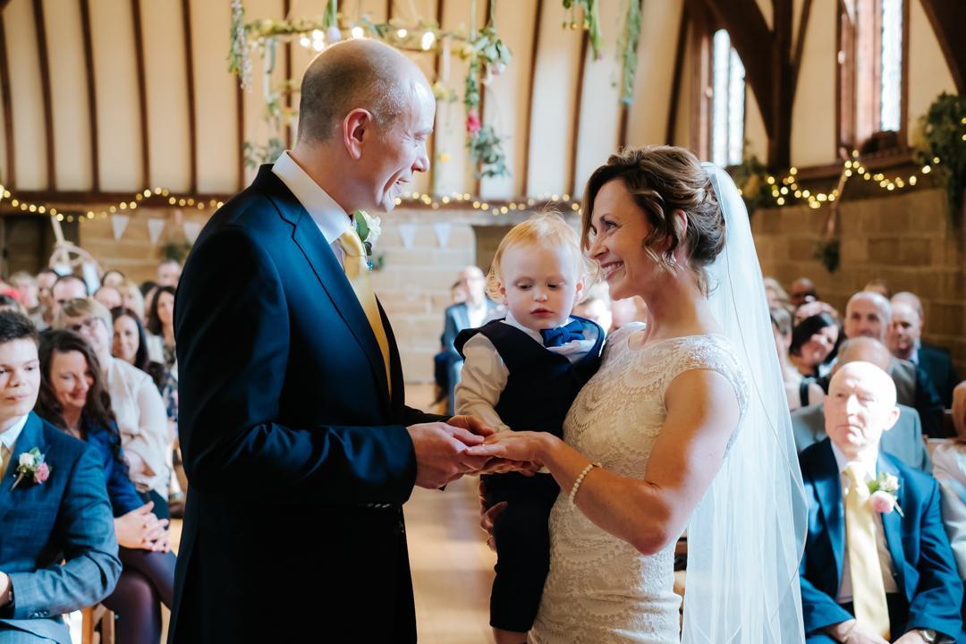 Groom puts wedding ring on his brides fingers whilst their son looks on at the Lord Lycester Hospital wedding venue