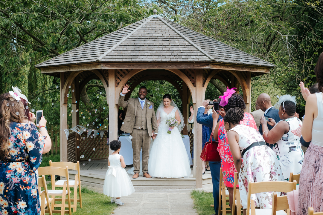 African groom and English bride stand in Worton Hall's wooden pagoda on the green in the gardens. The groom waves with glee having just been officially pronounced man and wife.