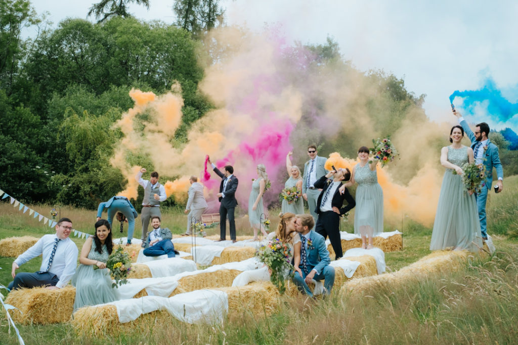 Bride and groom kiss whilst their ushers and bridesmaids have fun waving coloured smoke bombs in the air after their relaxed wedding ceremony at Hadsham farm weddings. my favourite kind of wedding to photograph