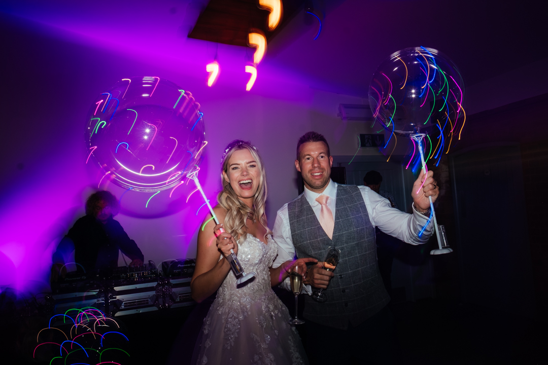 bride and groom stand together smiling on the dancefloor at their swallows nest barn wedding, holding light up balloons and glasses of champagne