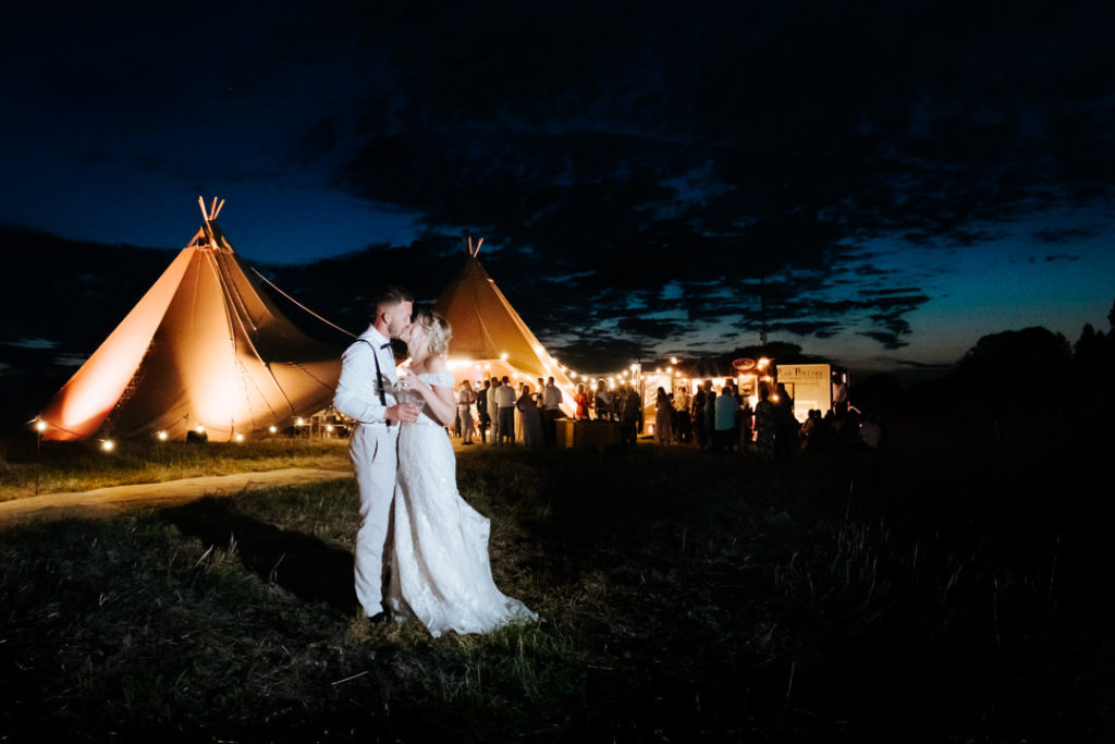 bride and groom stand kissing in front of thier wedding tipi lit up in the night sky 