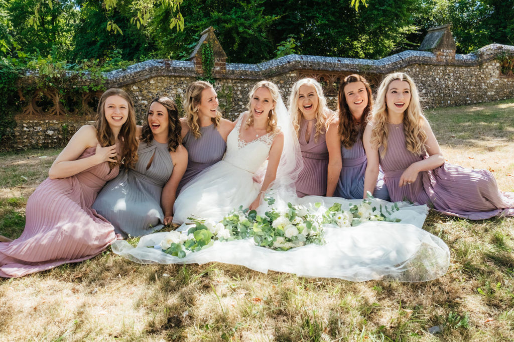 A bride and her bridesmaids sit on grass in dappled sunlight laughing and smiling together. Photographed by Charlie Flounders at the Barn at Avington wedding
