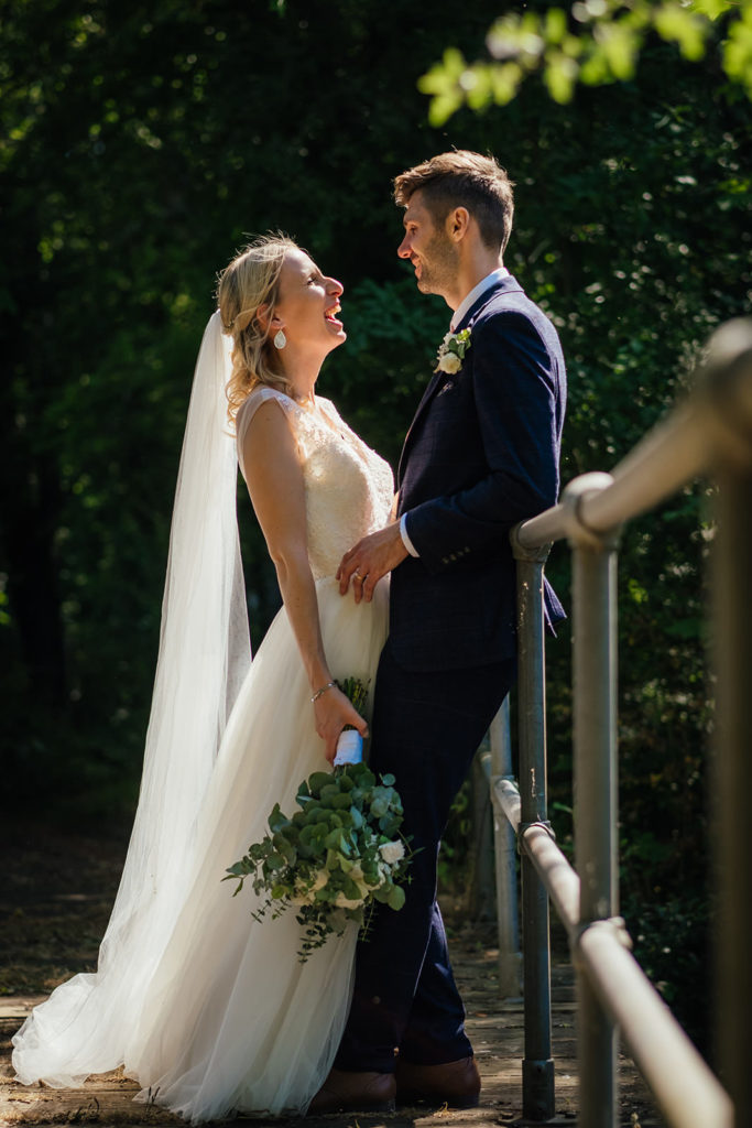 A bride and groom embrace on the bridge at The Barn at Avington whilst laughing together Photographed by Charlie Flounders at the Barn at Avington wedding