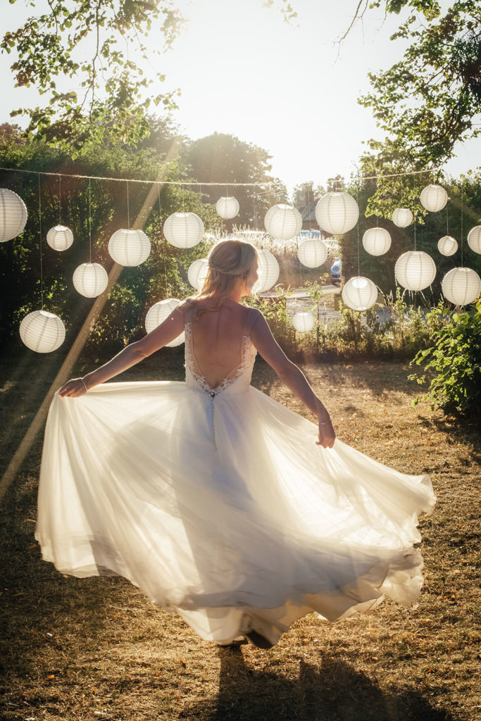 A bride swishes her floaty wedding dress around in beautiful golden evening sunlight with white paper lanterns in the background. Photographed by Charlie Flounders at the Barn at Avington