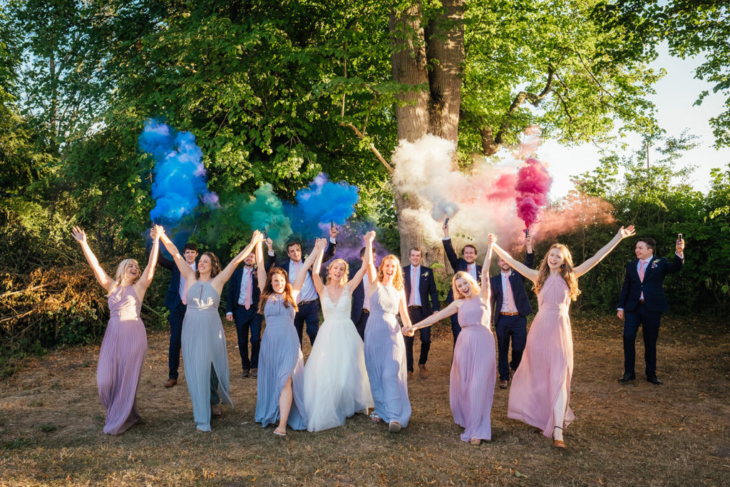 group of people at a wedding in pastel coloured dresses waving coloured smoke flares in the air. Photographed by Charlie Flounders at the Barn at Avington
