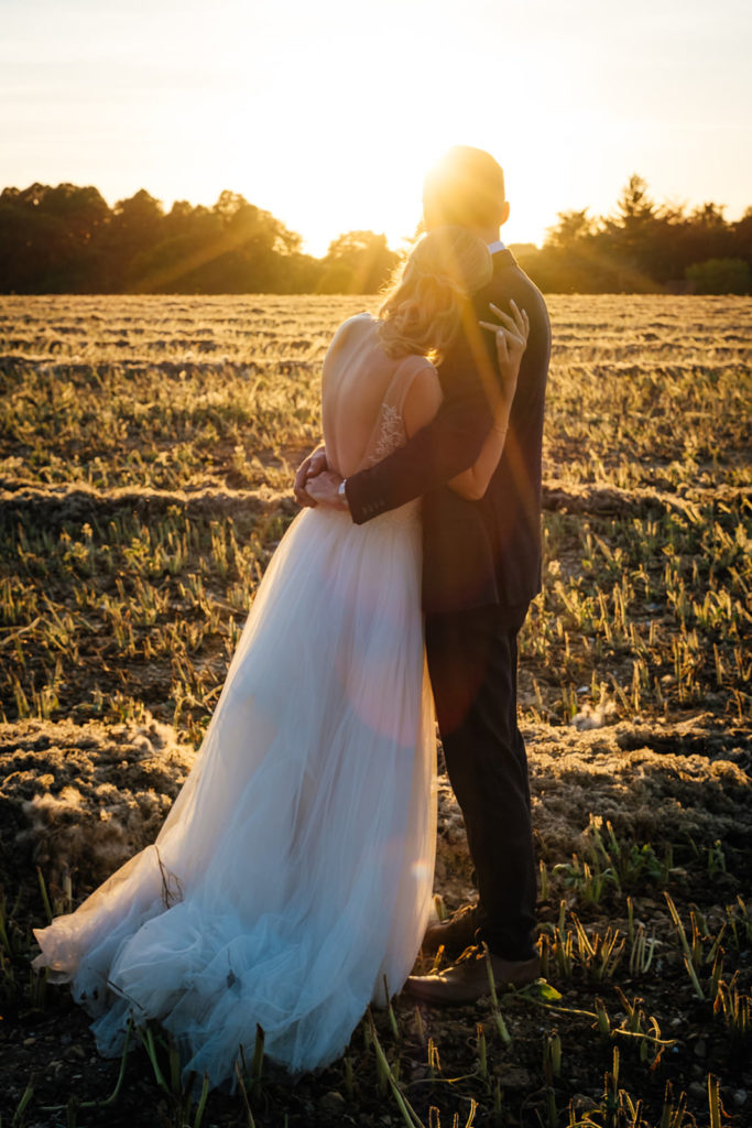 Bride and groom hug as they look on to beautiful golden sunlight across farm fields at the Barn at Avington. Photographed by Charlie Flounders